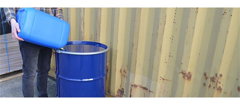 Someone pouring the contents of a blue jerry can into a blue steel drum against a shipping container